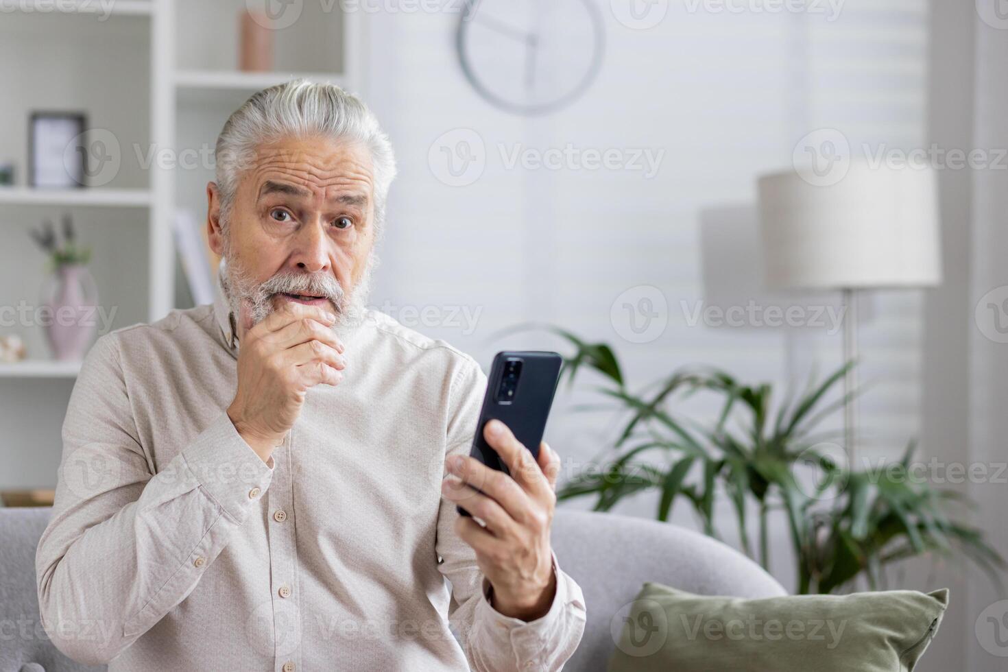 Close-up portrait of a worried senior pensioner man sitting at home on the sofa, holding a phone in his hand and looking at the camera in shock, holding his beard with his hand. photo