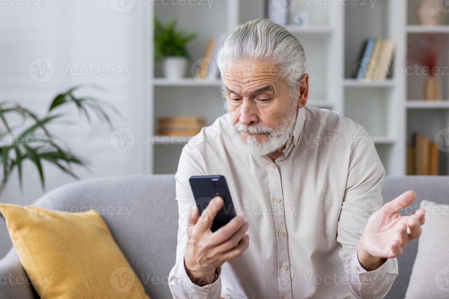 Elderly man sitting on couch using mobile phone, looking confused and expressing frustration with hand gesture. Home interior background with bookshelf and plants. photo