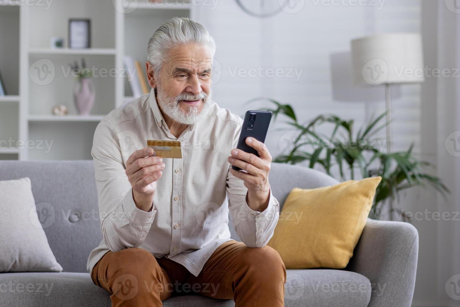 Smiling senior gray-haired man using mobile phone and credit card at home and sitting on sofa. photo