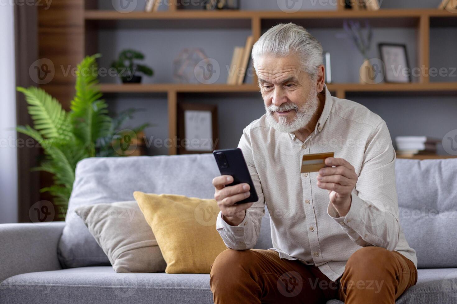 Gray-haired scared man uses credit card through mobile app, holds phone, sits on sofa at home. photo