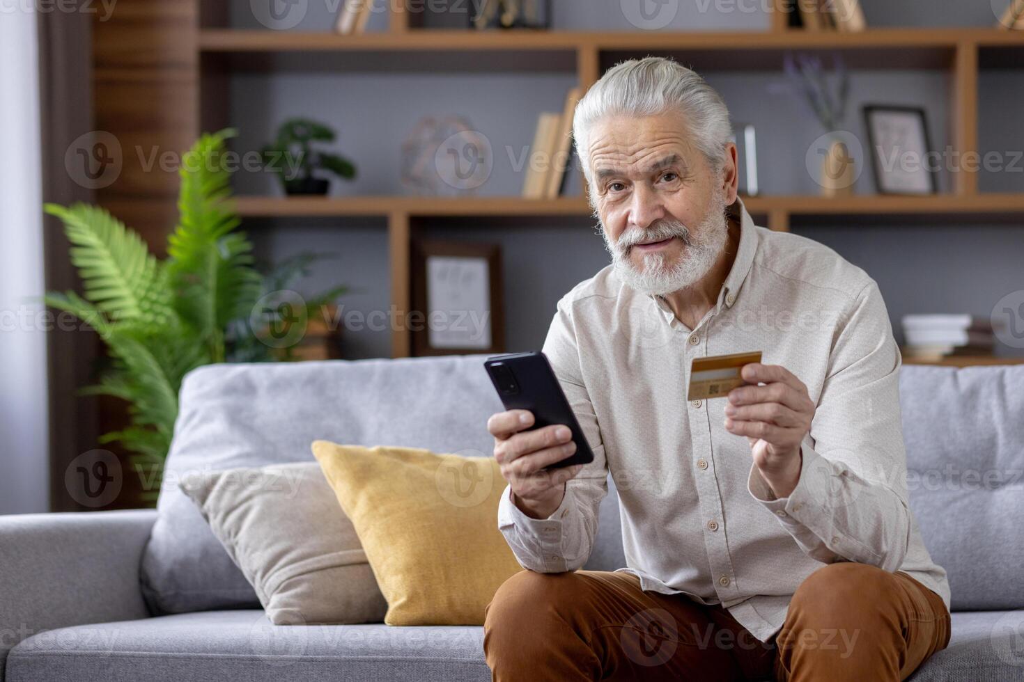 Portrait of a smiling senior man at home, sitting on the couch and looking at the camera. holds a credit card and mobile phone. photo
