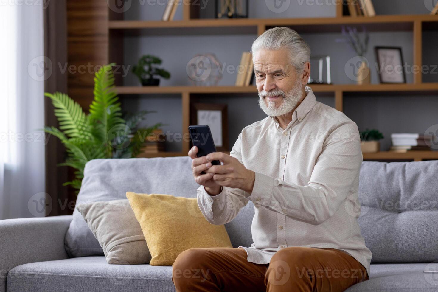Senior man sitting on a couch in a cozy living room, using a smartphone and smiling. Concept of technology, communication, and leisure. photo