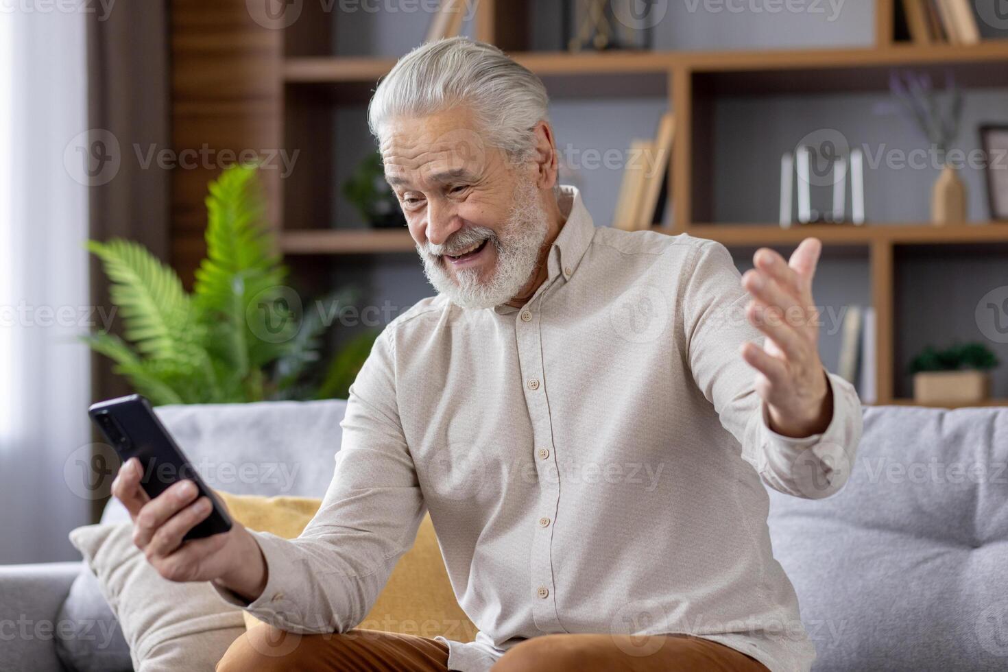 Smiling senior gray-haired man grandfather talking on call on the phone, sitting on the sofa at home and happy with the news. photo