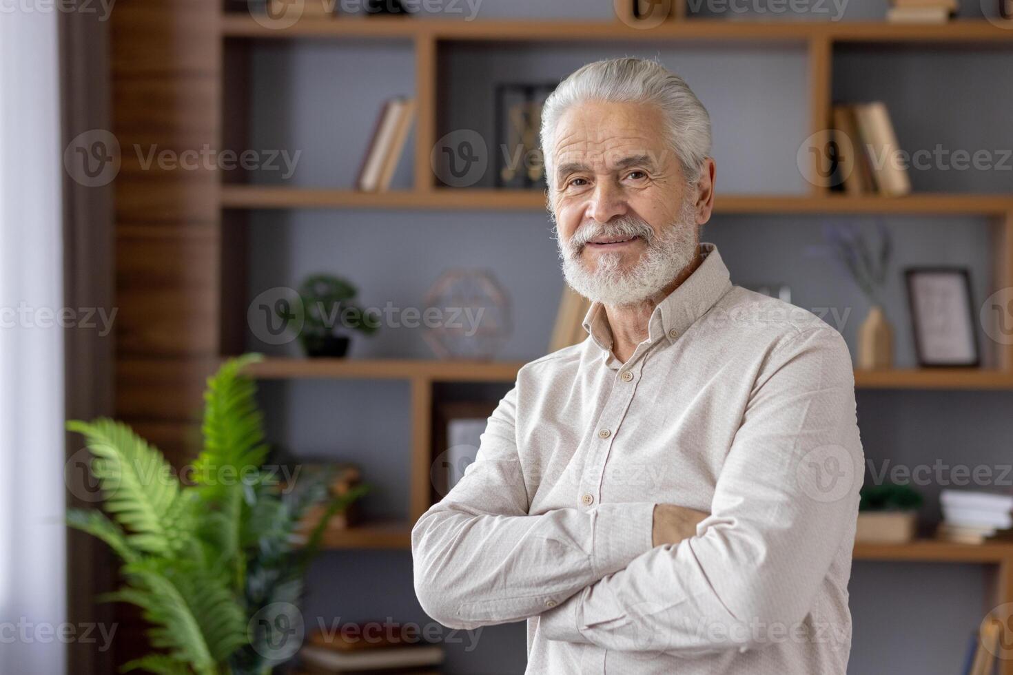 Portrait of a scary gray-haired man standing in a home office, crossing his arms on his chest and looking confidently and smiling at the camera. photo