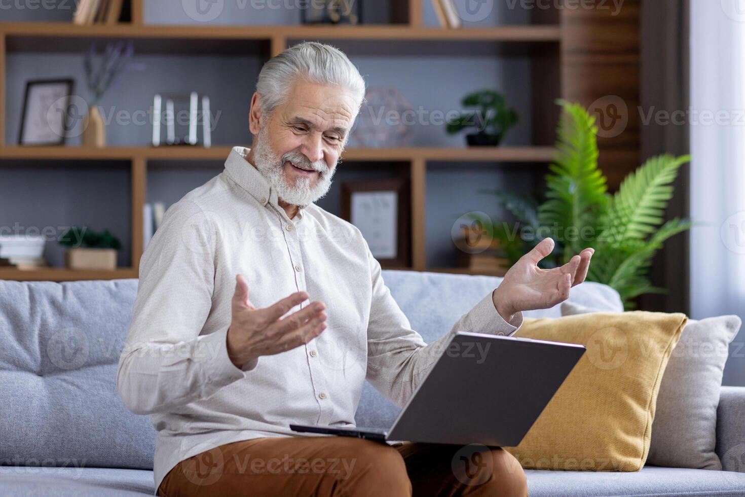 Gray-haired old smiling man is having a call on a laptop, sitting on the sofa at home and talking remotely. photo
