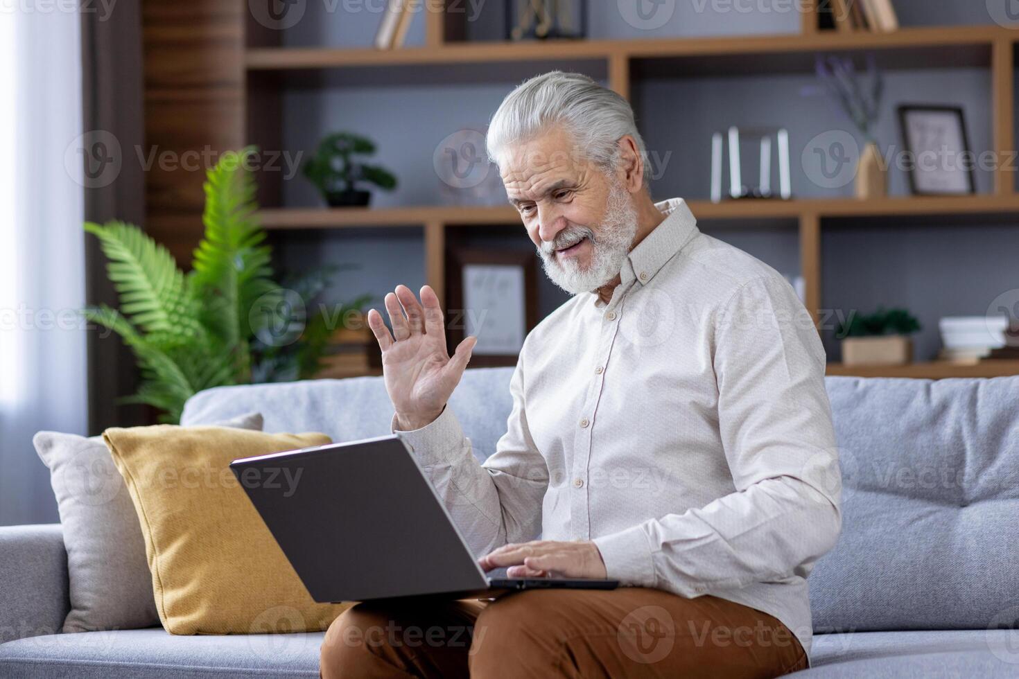 Smiling gray-haired senior man sitting at home on sofa with laptop and talking on call to camera, waving and greeting. photo
