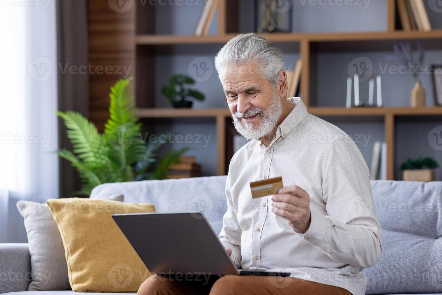Senior gray-haired man sitting at home on the sofa and using a laptop, holding a credit card and conducting online financial transactions. photo