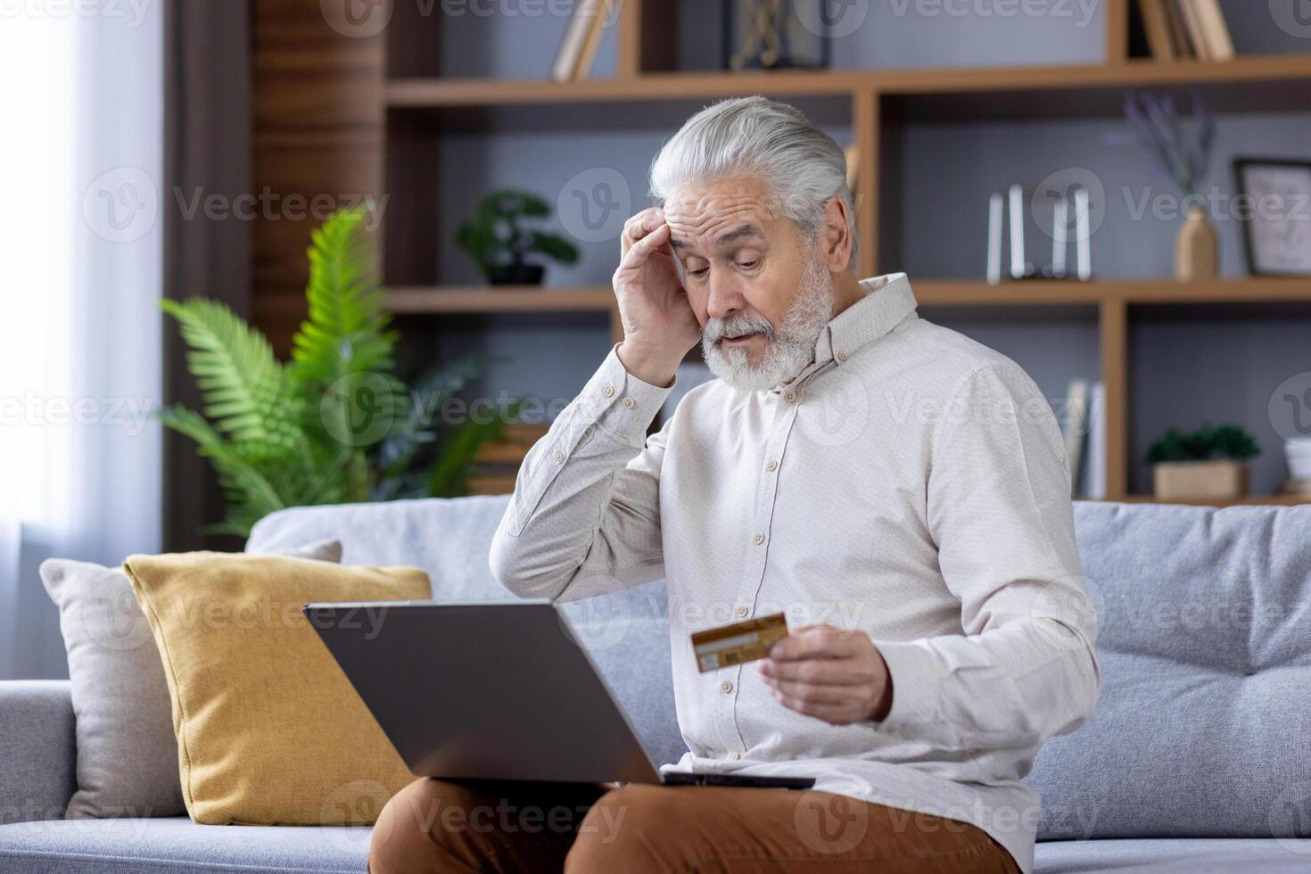 Worried senior gray-haired man sitting on sofa at home with laptop, holding head, having credit card problem. photo