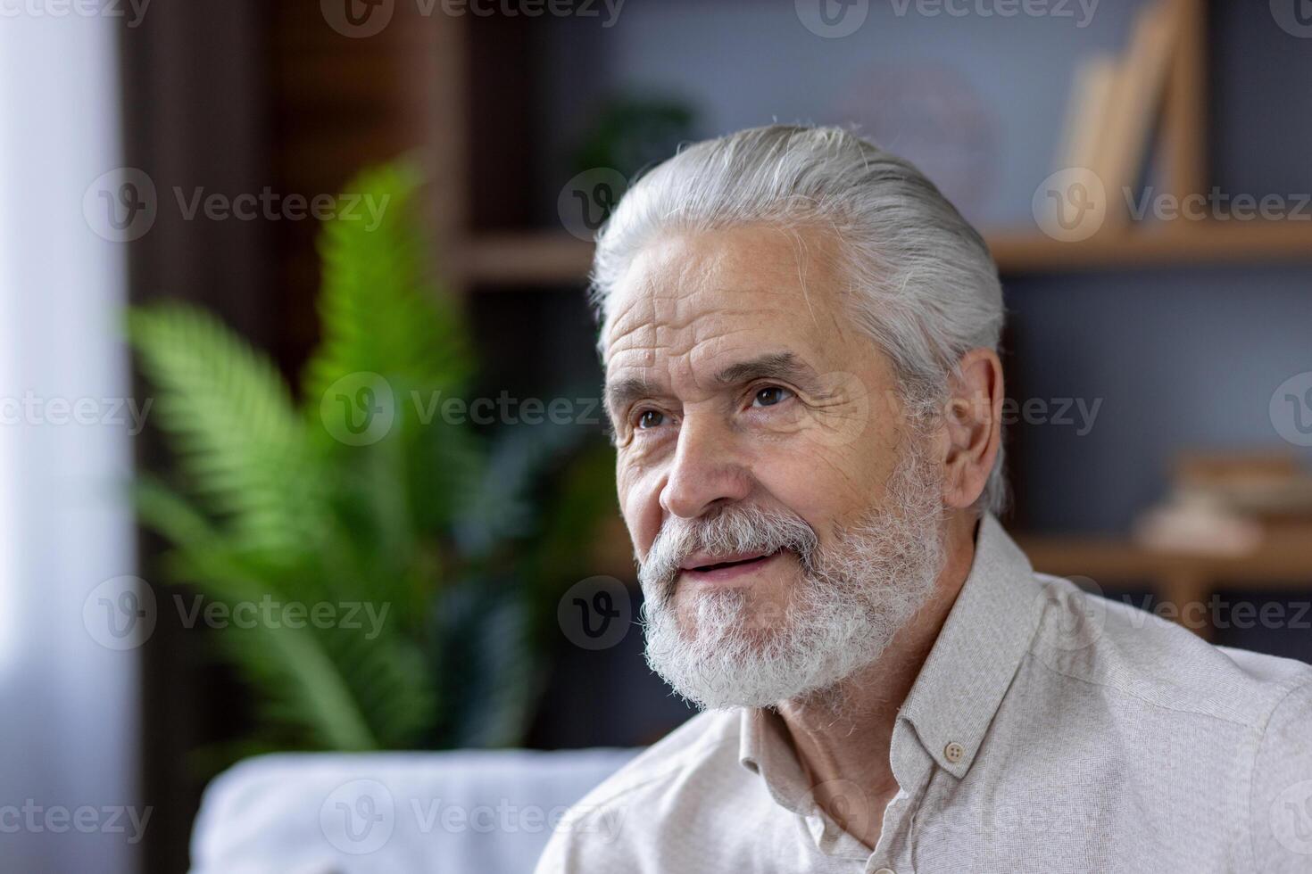 Close-up photo of a smiling gray-haired and bearded senior man at home, thoughtfully and thoughtfully looking ahead.