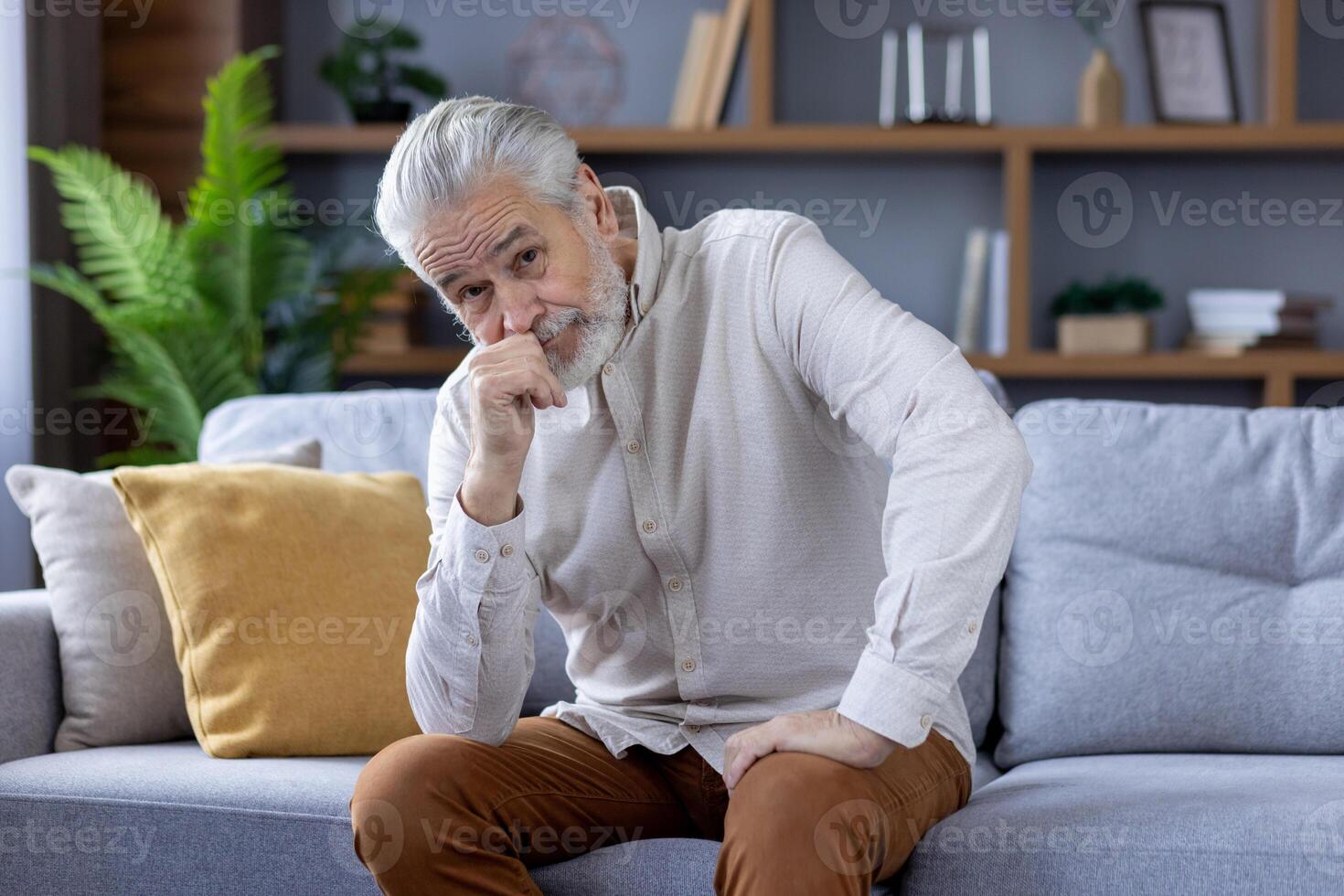 Portrait of a serious and pensive senior man sitting on the couch, leaning his head on his hand and looking sadly into the camera. photo