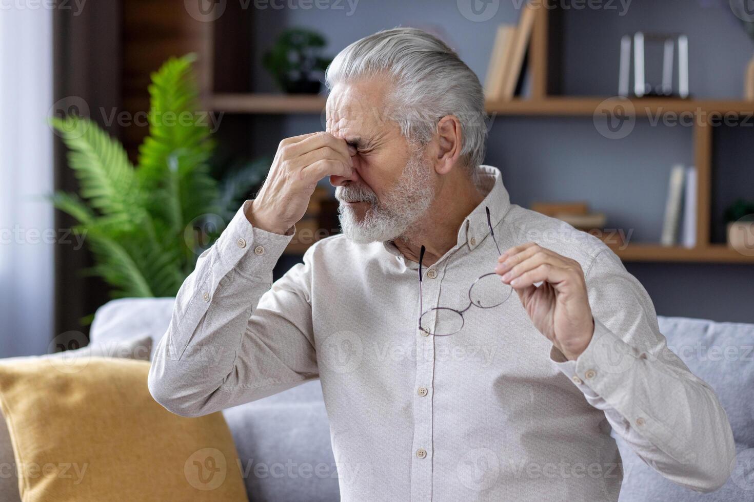 Tired gray-haired man sitting on the sofa at home, holding glasses in his hand and rubbing his eyes with his hand, resting. photo