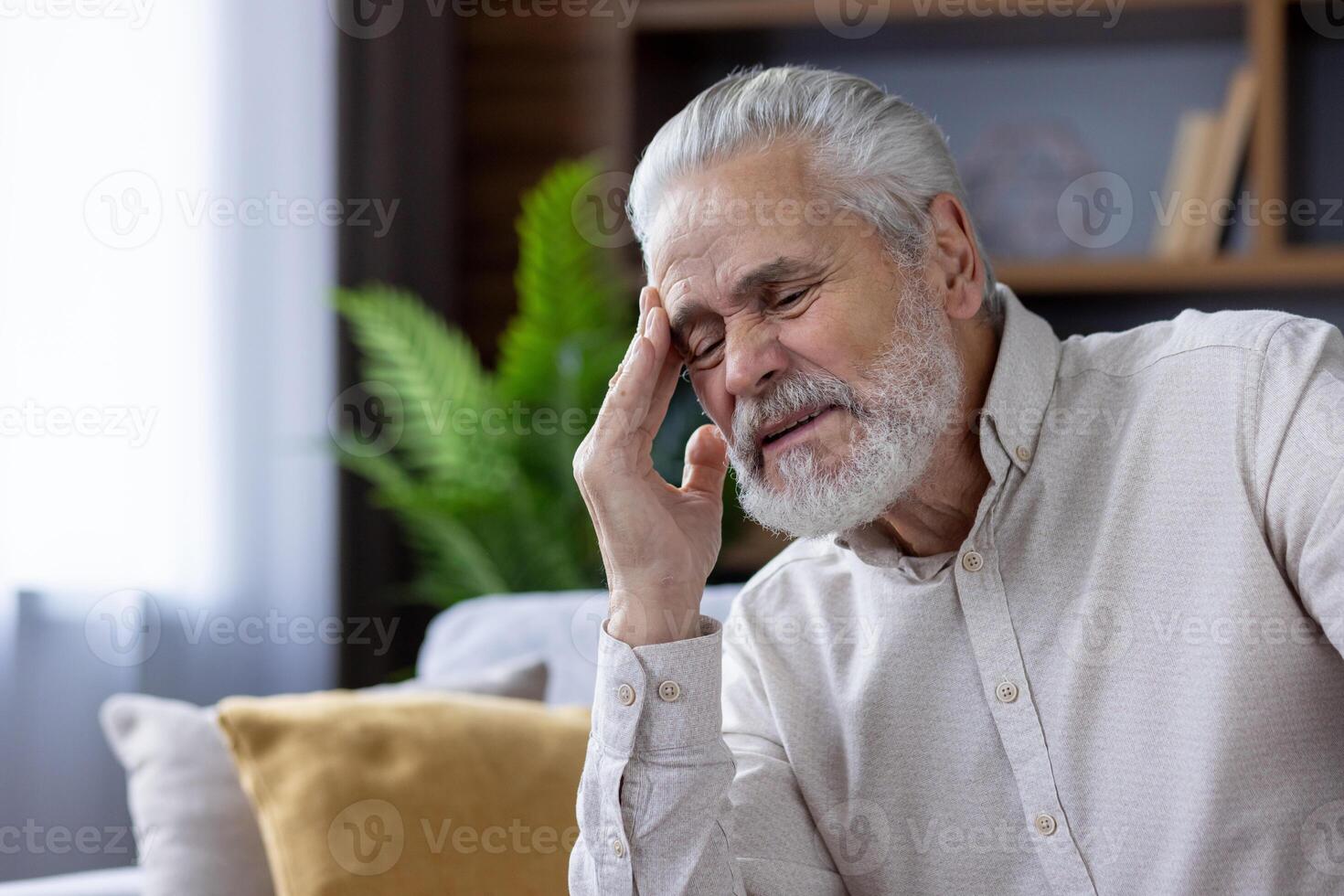 Close-up photo of a tired and sick old gray-haired man sitting at home on the sofa and holding his head with his hand, closed his eyes and is in pain.