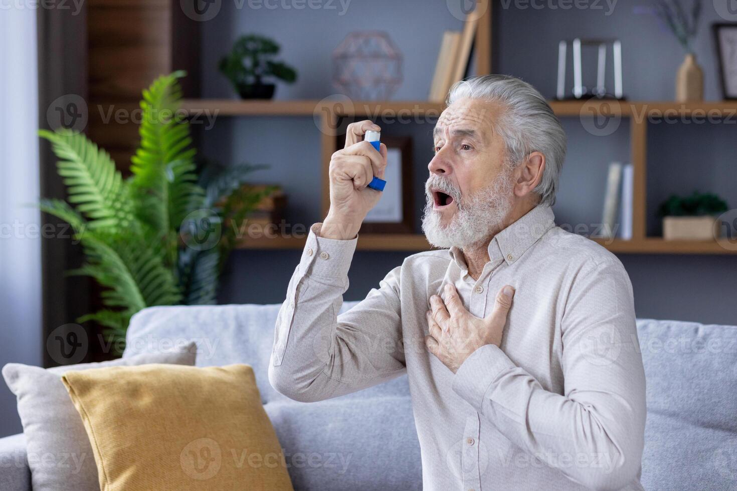 An elderly gray-haired man suffers from asthma. He sits at home on the couch and uses an inhaler. photo