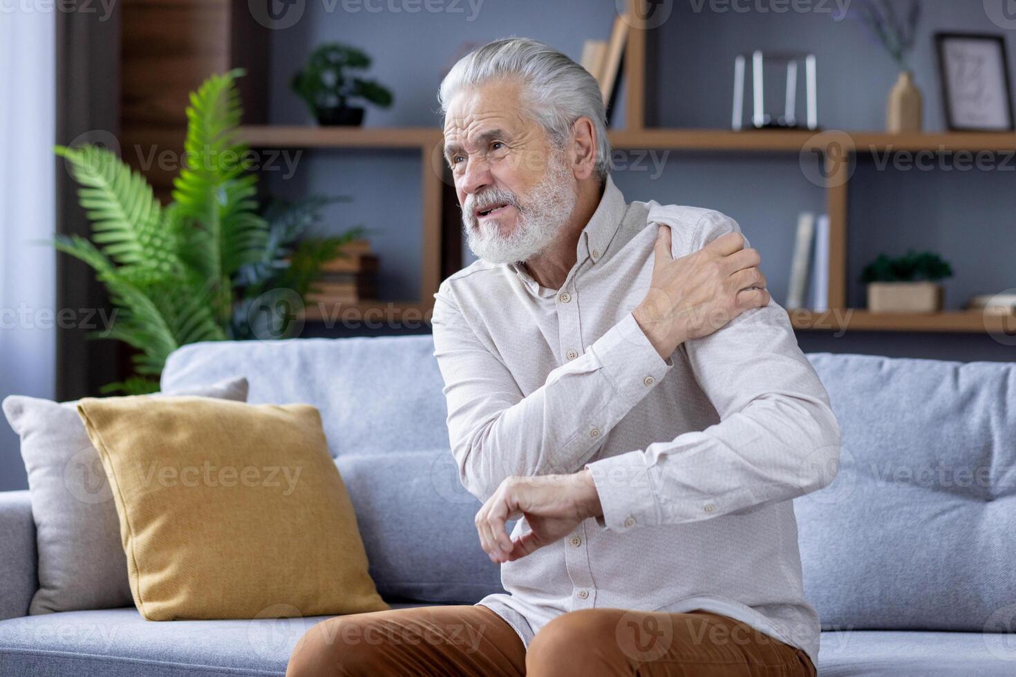 Senior gray-haired man sitting on sofa at home and holding arm above shoulder, suffering from pain and sprain. photo