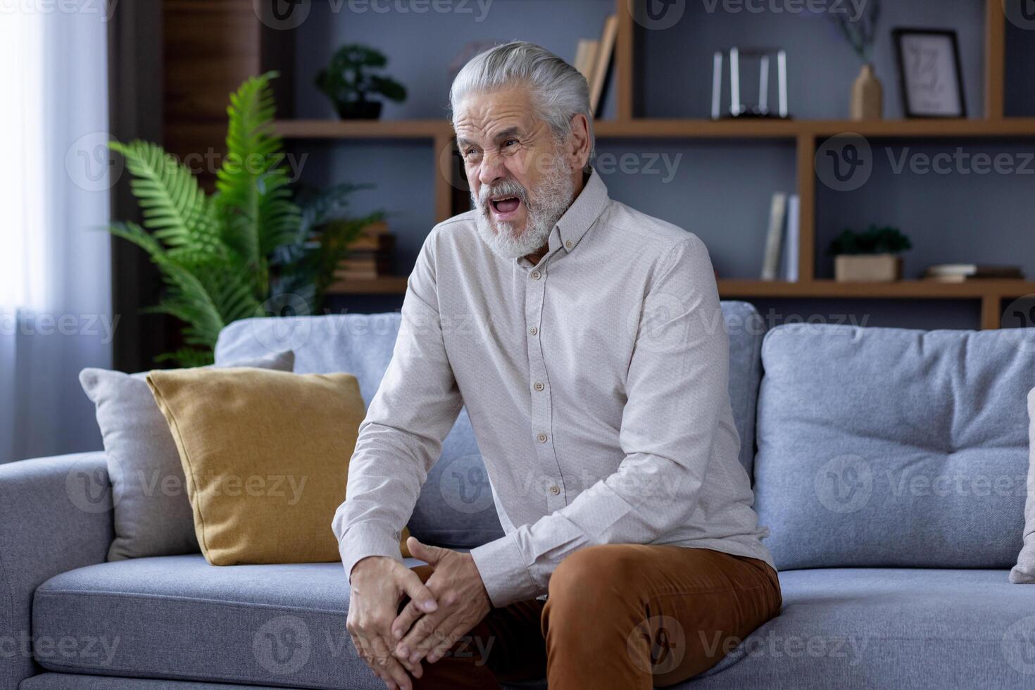 Grimacing from severe pain, an older gray-haired man sits on the sofa at home and holds his knee with his hand. photo