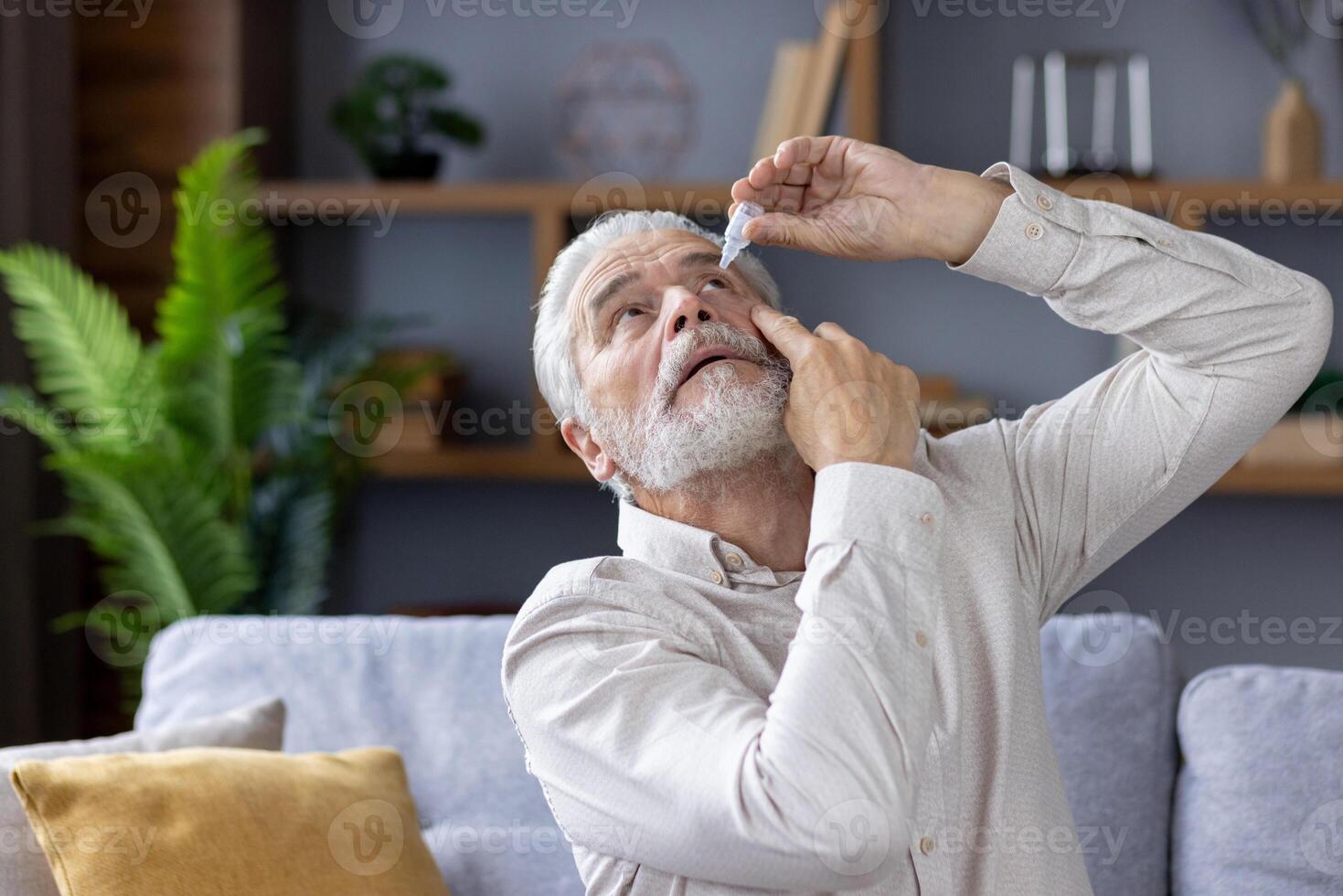 An elderly retired man is at home, sitting on the sofa and instilling medicine in his eye for inflammation and fatigue. The photo is close.