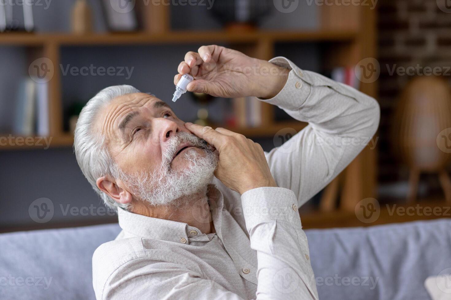 Close-up photo of an older gray-faced man who drips eye drops with medicine for pain and fatigue, sitting on the couch at home.