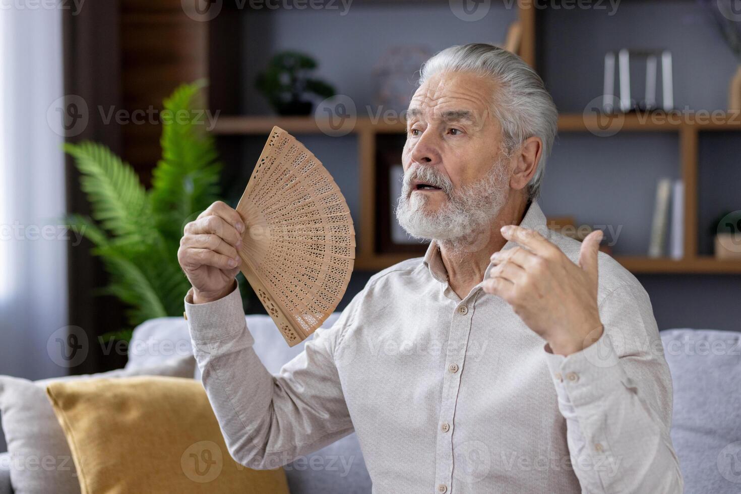 Close-up photo of a gray-haired senior man sitting on the sofa at home and suffering from insomnia, holding a fan in his hand and waving in his face.