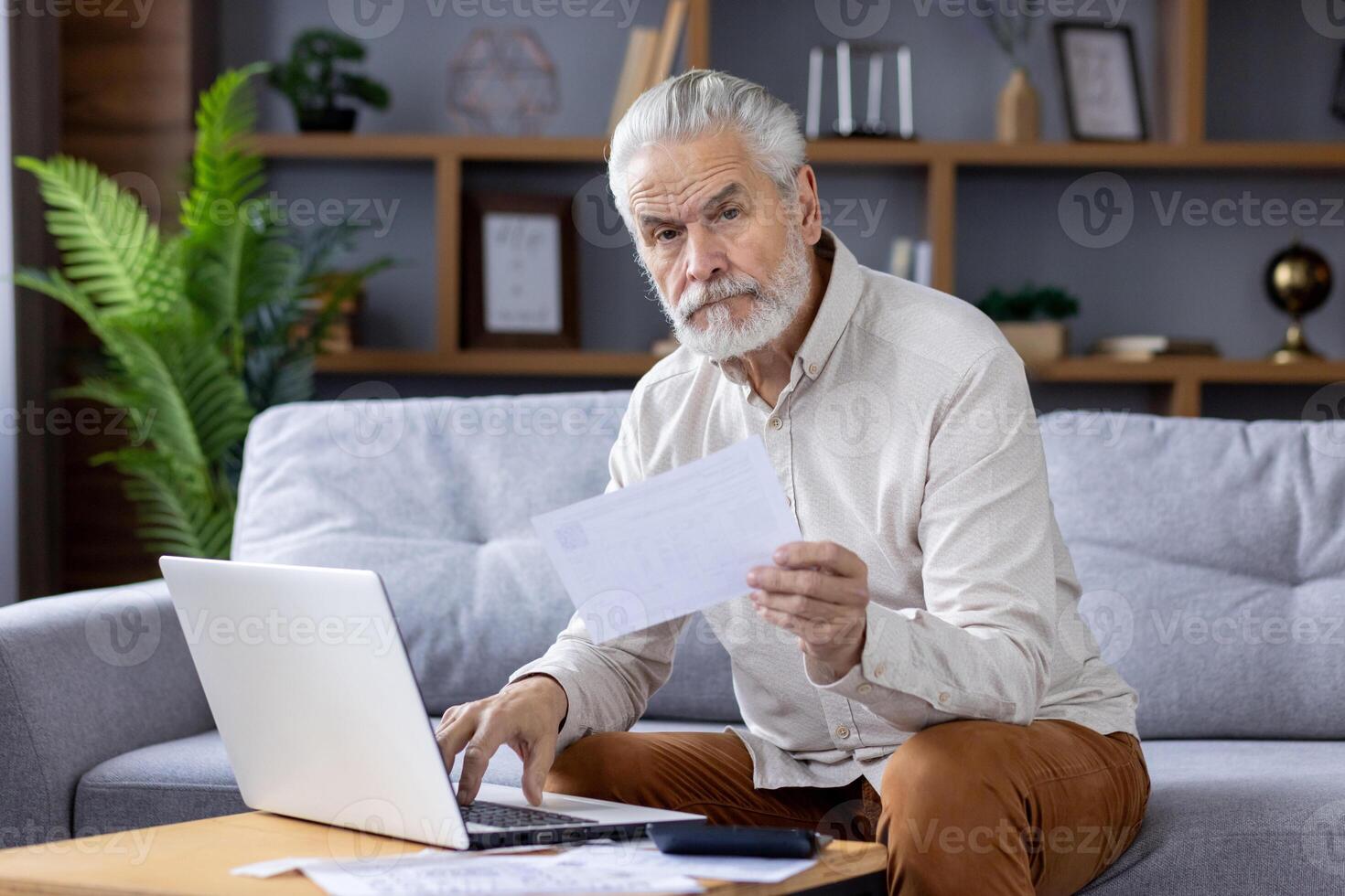 Senior gray-haired man sits on the sofa at home and works on a laptop, holds bills and documents in his hands, looks seriously into the camera. photo