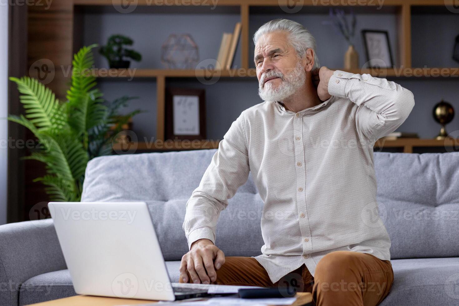 Tired senior gray-haired man sits at home on the sofa in front of the table with a laptop and holds his neck with his head, doing asana, resting. photo