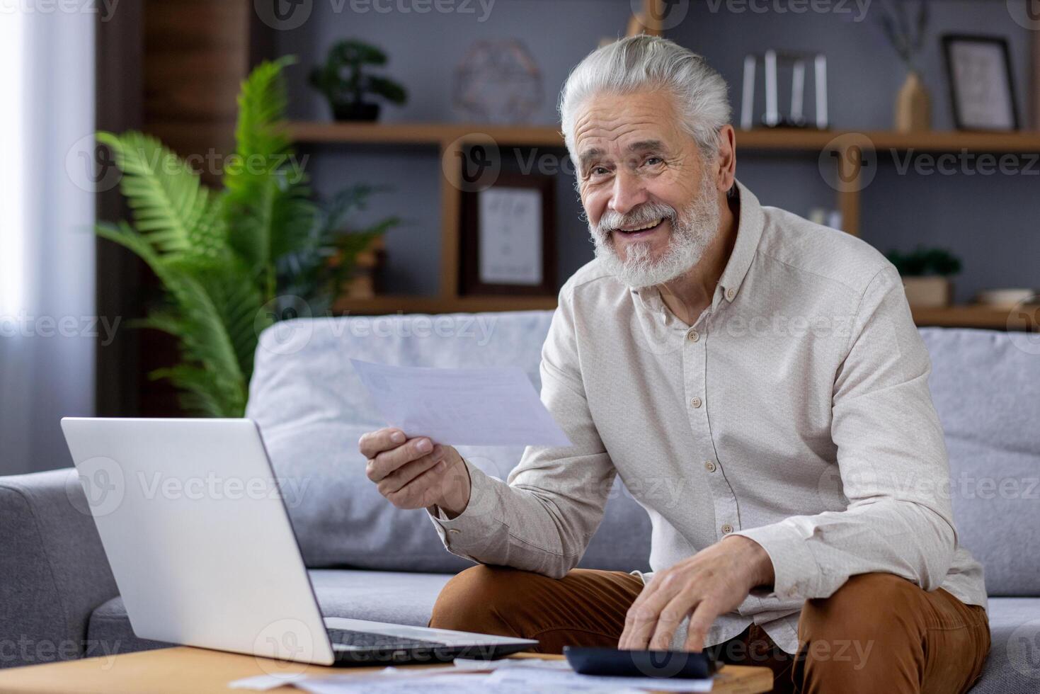 Portrait of smiling senior gray-haired man sitting on sofa at home and checking bills and documents, using laptop and calculator, looking at camera. photo