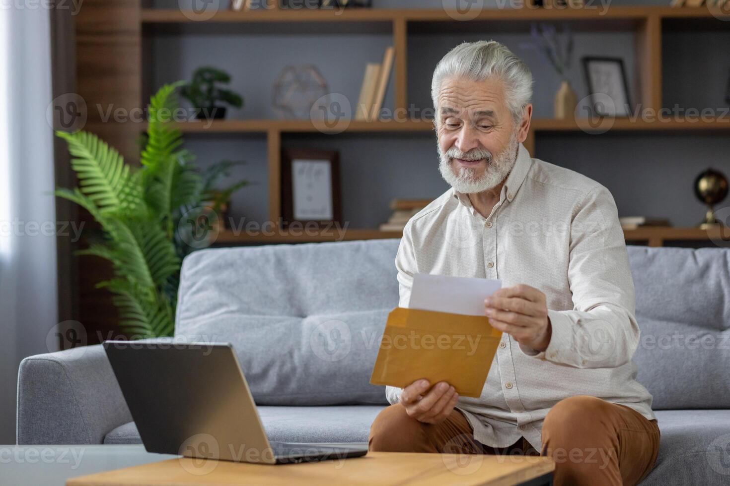 Senior gray-haired smiling man sits on a sofa and opens an envelope with a letter. Reads the received message, bill, invitation. photo