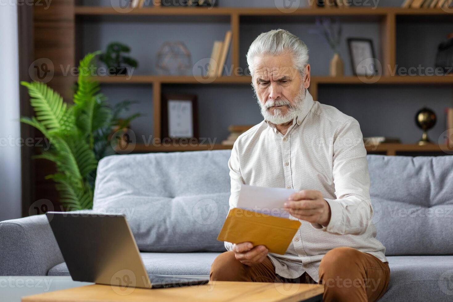 Worried and upset senior man sitting on sofa at home and opening envelope with letter, received bills, bad news, medical results. photo