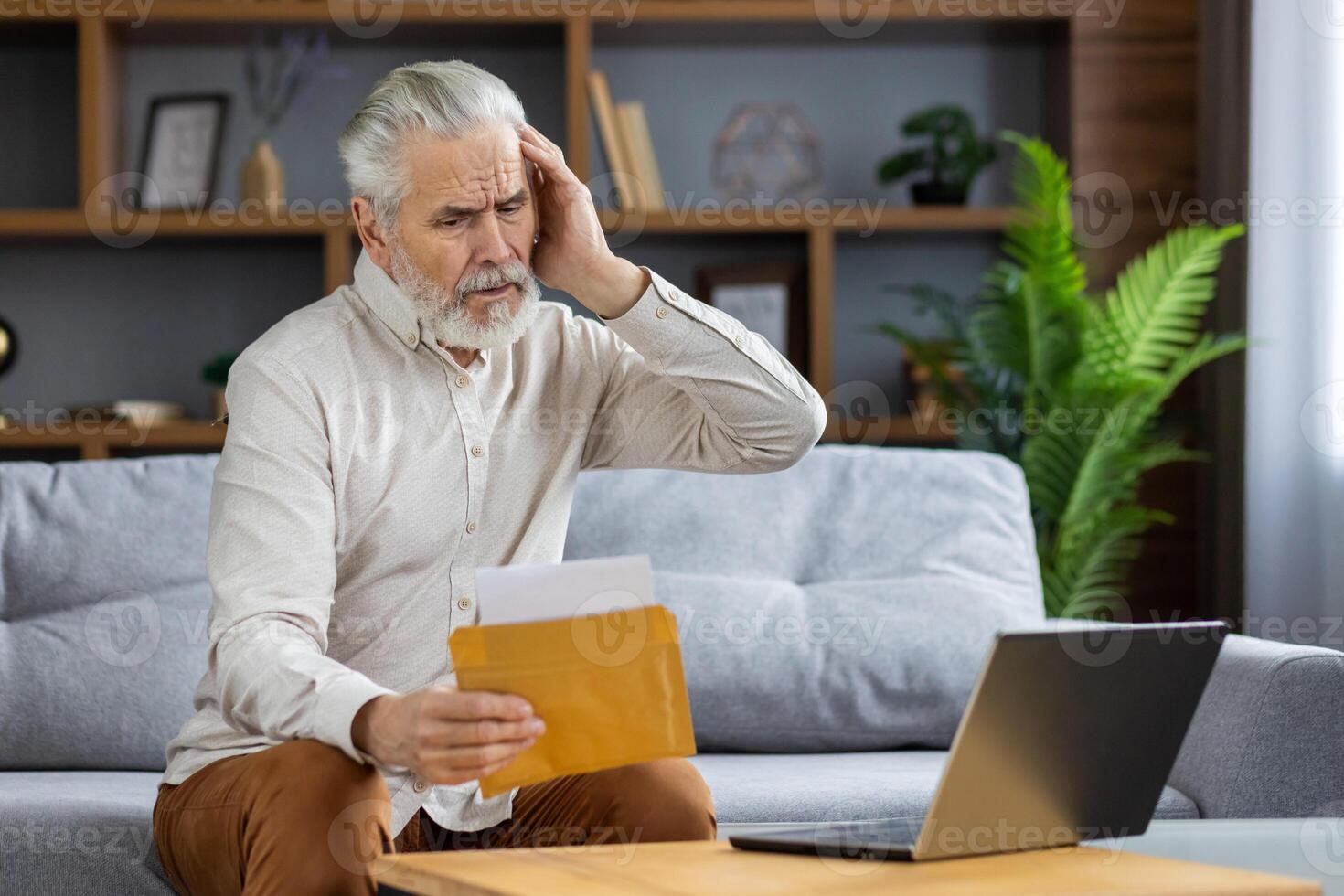 Upset senior gray-haired man sitting at home on sofa in front of laptop and holding envelope with letter with bad news. Worriedly holding his head. photo