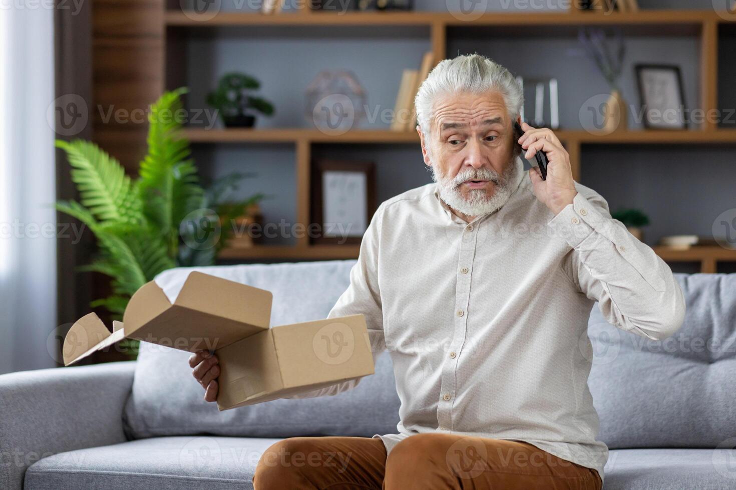 An older gray-haired man sits at home on the sofa and angrily talks on the phone, stubbornly holding in his hands an open package delivered by mail. photo