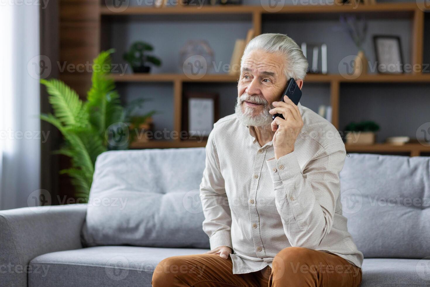 A gray-haired senior man is sitting on the sofa at home and talking on the mobile phone. photo