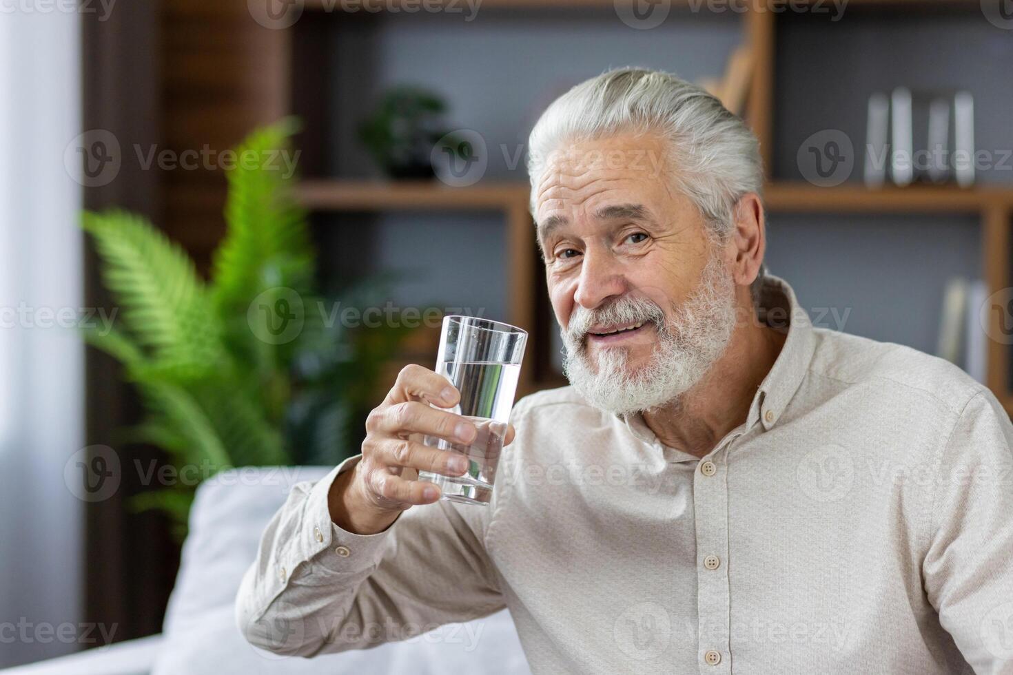 Close-up portrait of an elderly gray-haired man sitting at home on the couch holding a glass of clean water, leading a healthy lifestyle, pointing at the camera and smiling. photo