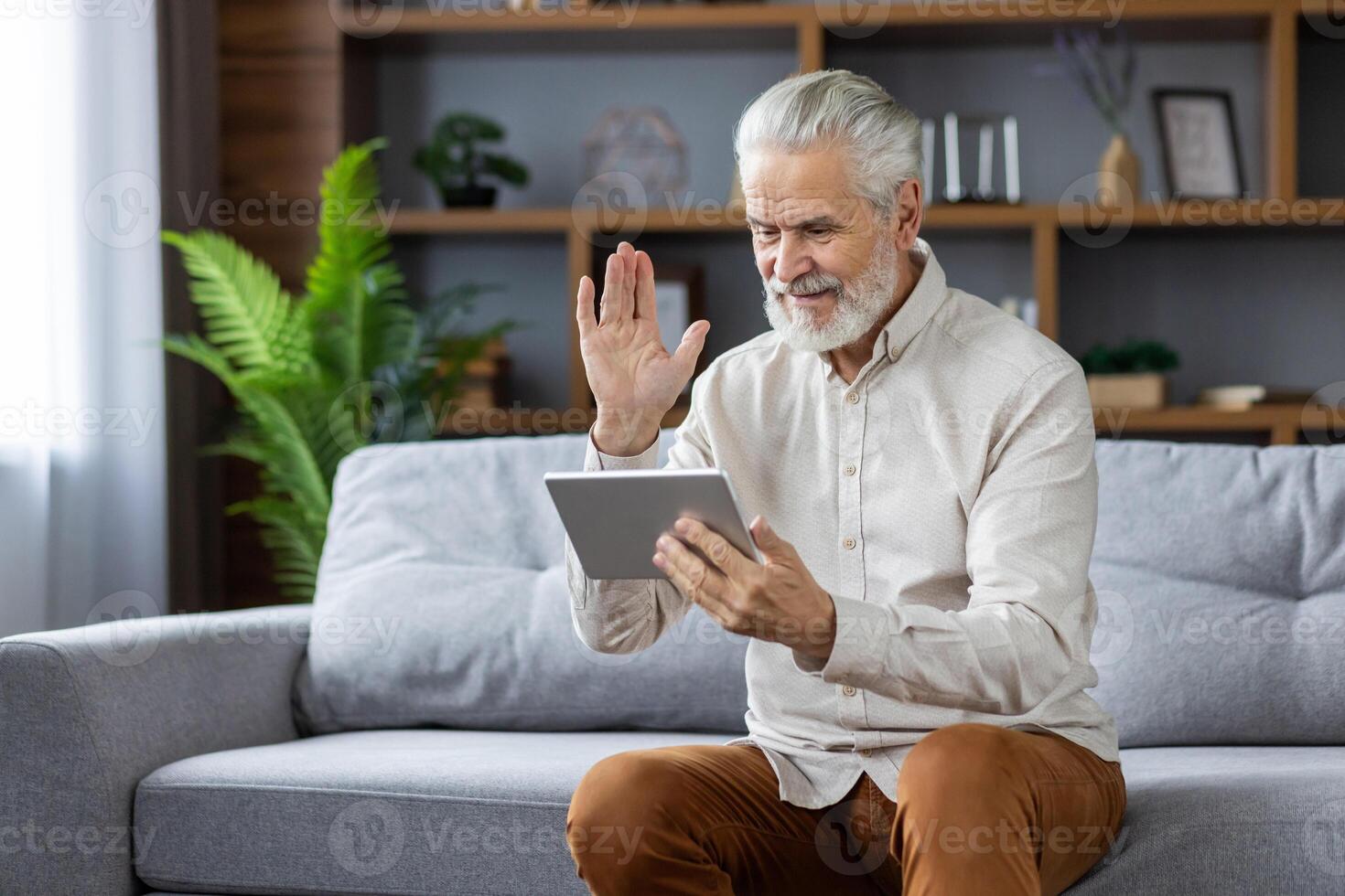 Gray-haired senior man communicating online via call from tablet, greeting and waving, sitting on sofa at home. photo