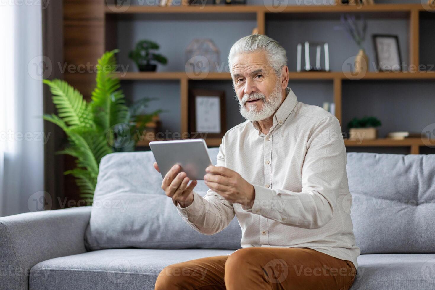 Senior gray-haired man sitting on the sofa at home, using a tablet, talking on a call and looking at the camera. photo