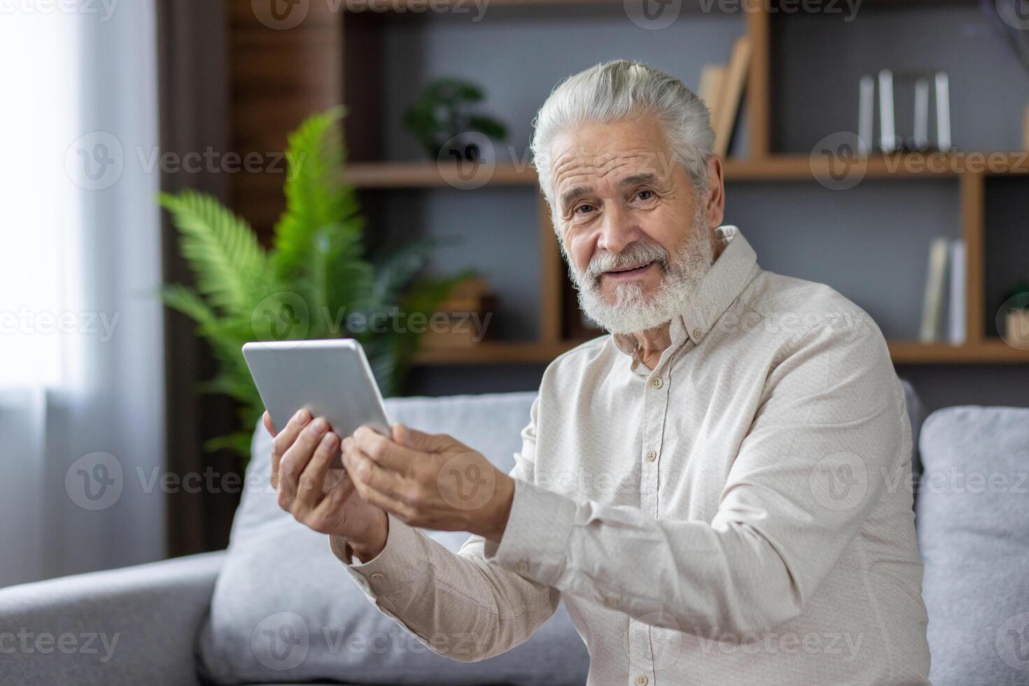 Close-up portrait of an older gray-haired man sitting on the couch at home, holding a tablet and smiling at the camera. photo