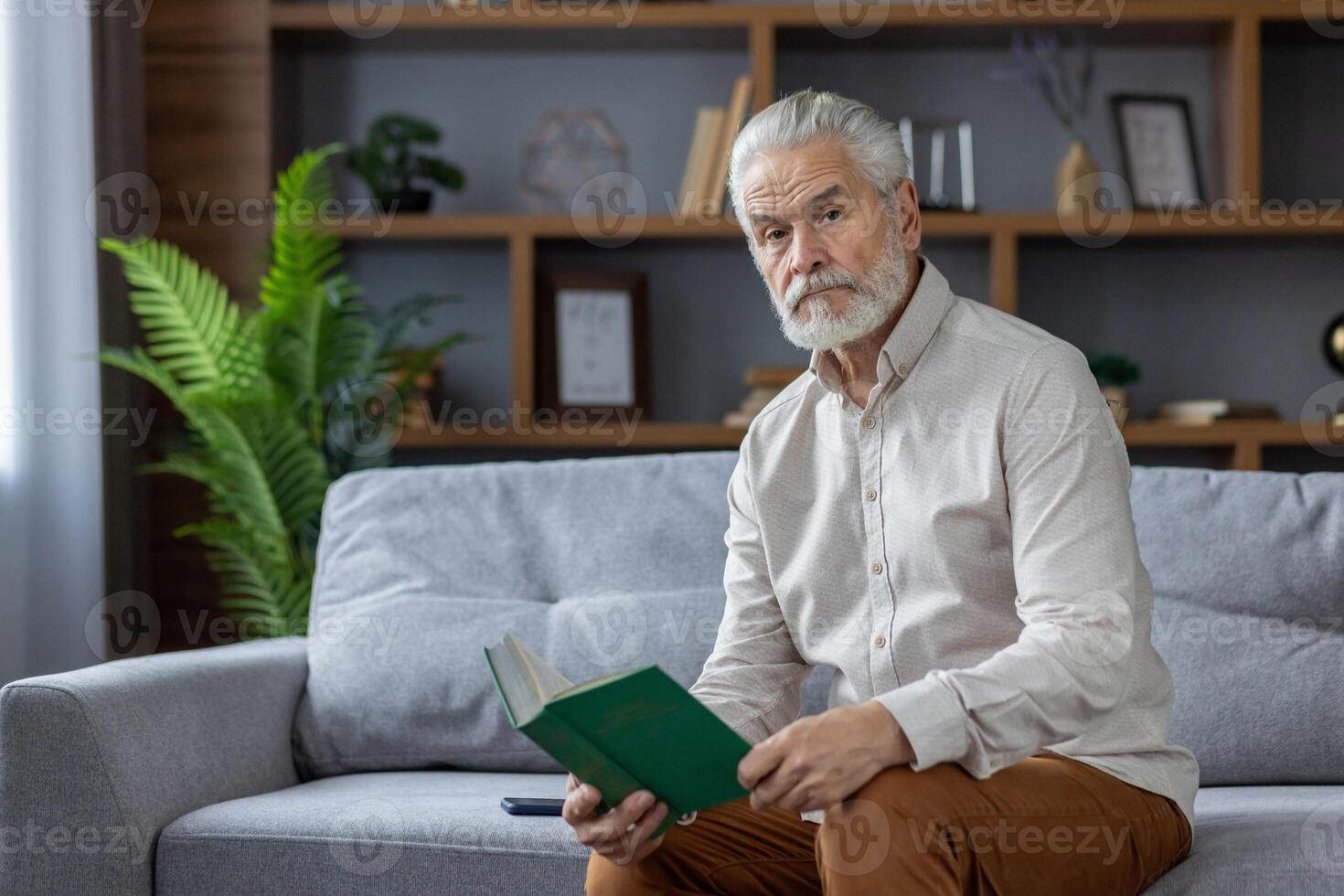 Portrait of a gray-haired senior man sitting at home on the couch, holding a book in his hands and looking seriously into the camera. photo