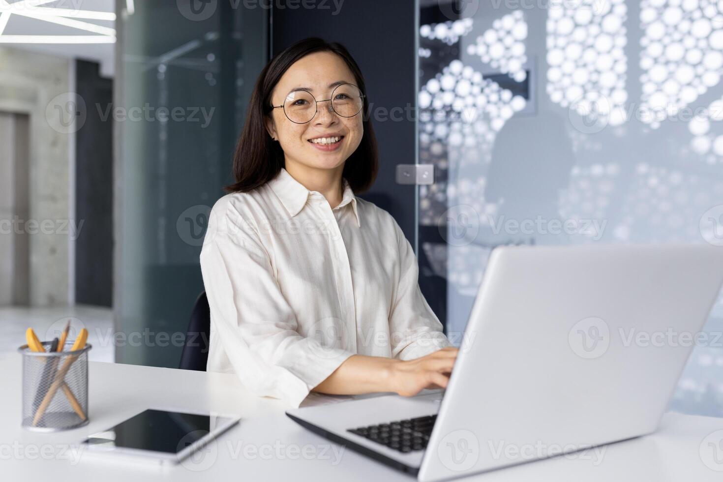 Young beautiful asian successful woman working inside modern office building, using laptop at workplace, businesswoman smiling and looking at camera, female boss happy with achievement results. photo