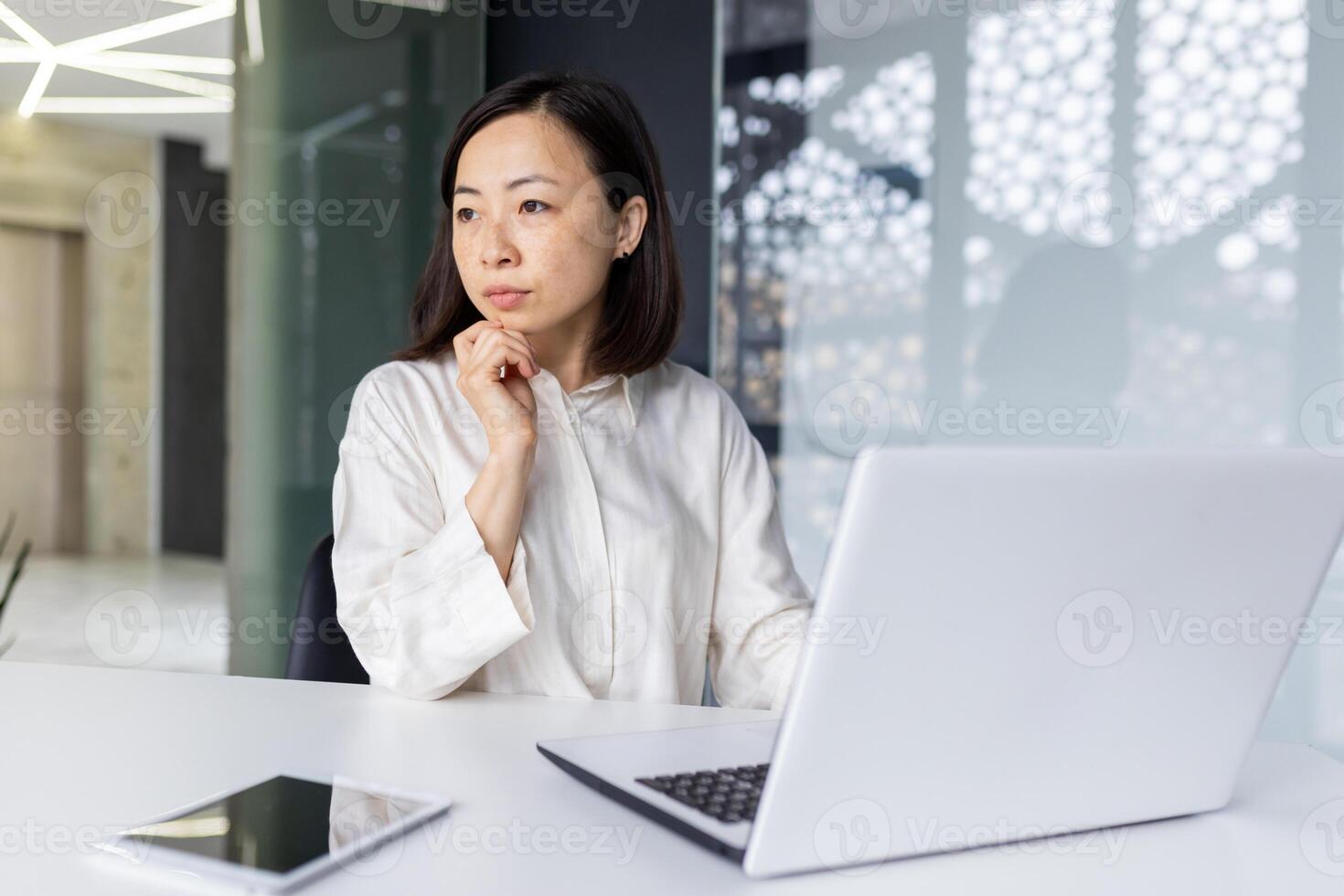 Photo of a successful and serious Asian business woman sitting in the office at a desk, working on a laptop and looking thoughtfully to the side.