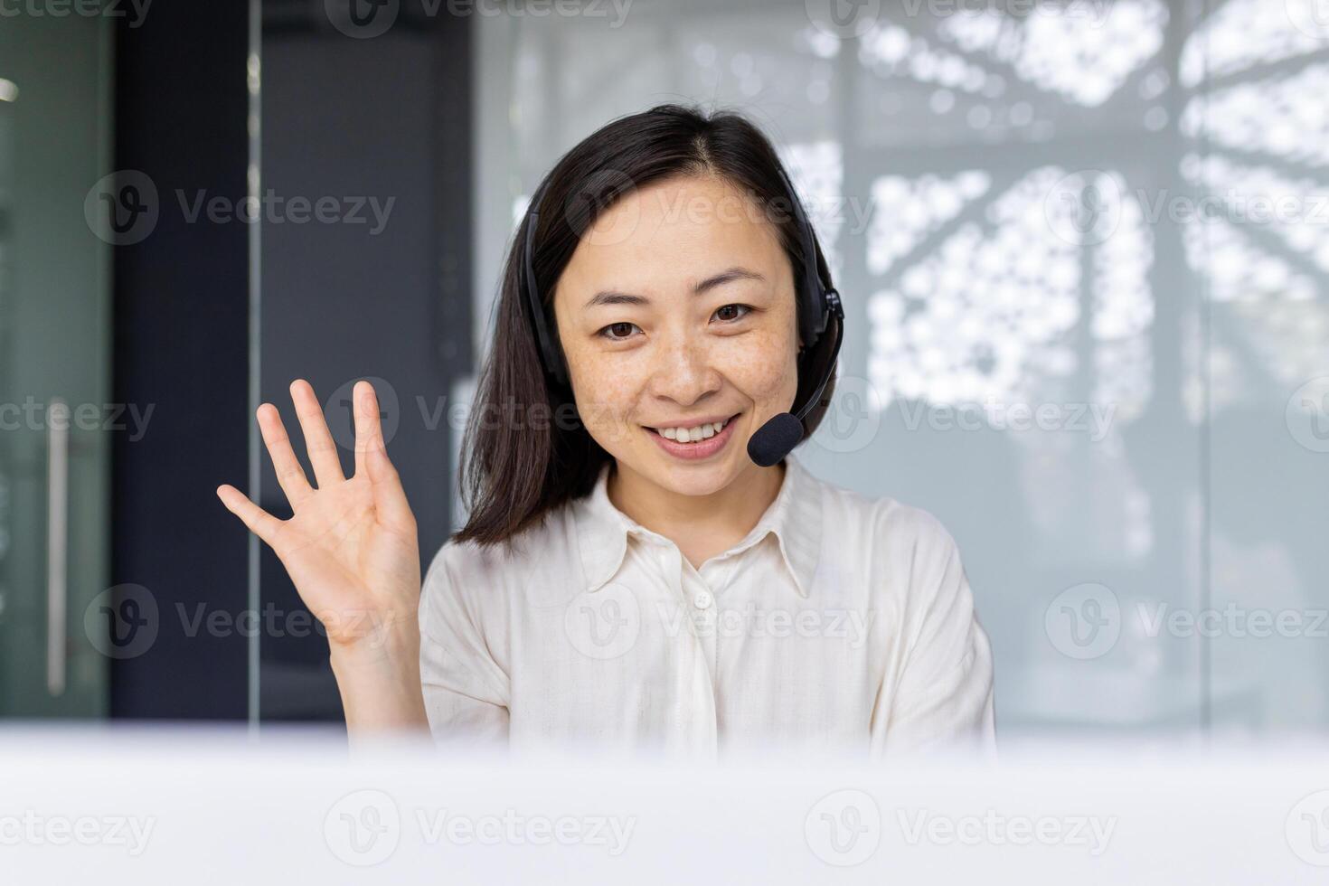 Professional Asian businesswoman in office with headset smiling and waving hand, symbolizing approachability and customer service. photo