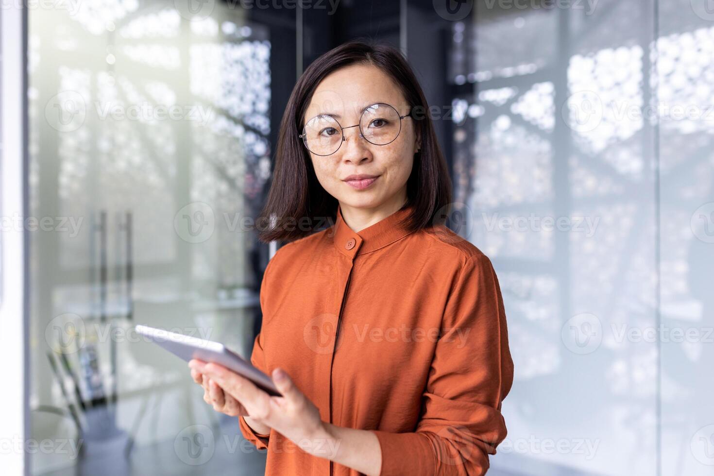 Portrait of a young Asian woman programmer, developer, tester in glasses standing in the office, holding a tablet, working online and looking, posing at the camera. photo