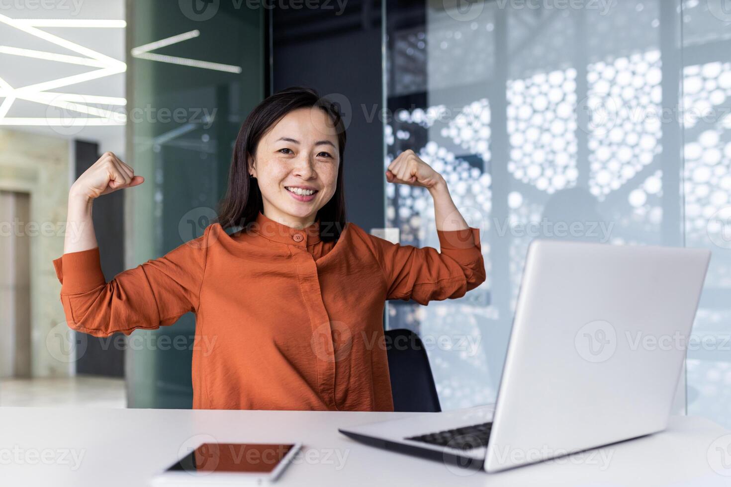 A young Asian business woman working in the office using a laptop, confidently and smilingly looking at the camera and showing a power gesture with her hands. Successfully completed work, project. photo
