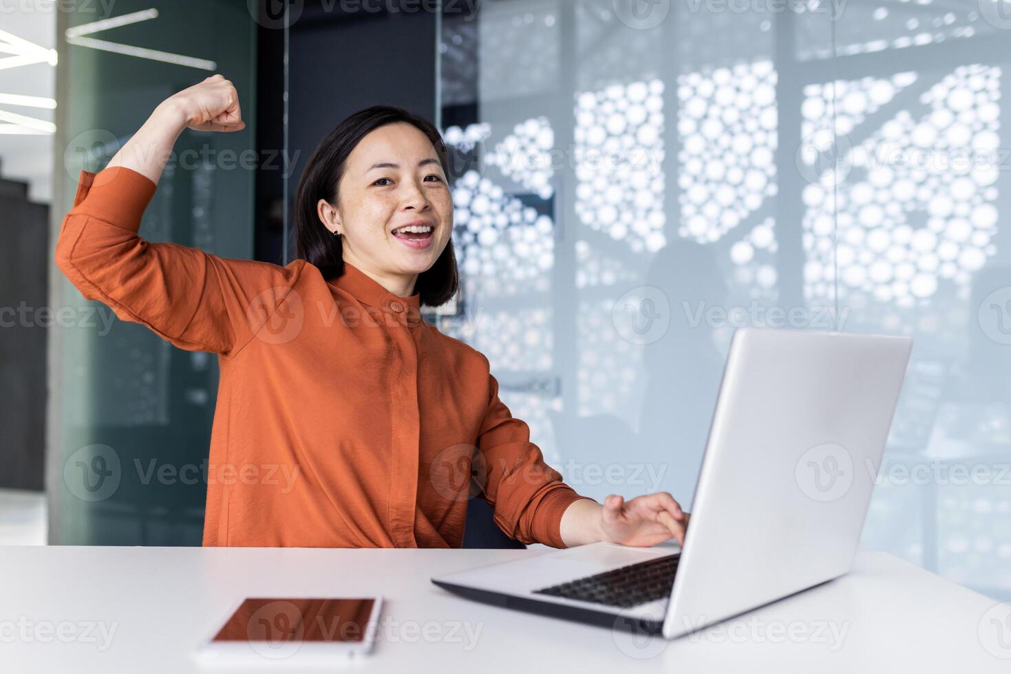 A young Asian woman student studying using a laptop in a co-working space. Satisfied with the test result, the examinee shows a hand gesture of strength and success. Smiling, she looks at the camera. photo
