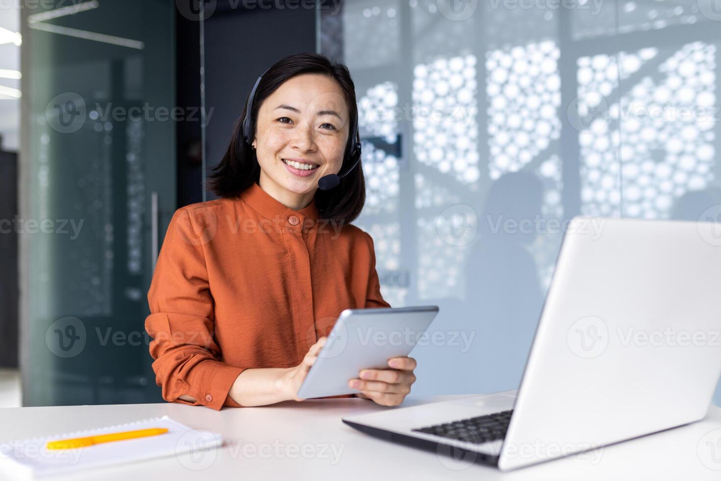 Portrait of successful satisfied female online customer service support worker, Asian business woman looking at camera, holding tablet computer, using headset and laptop for call. photo