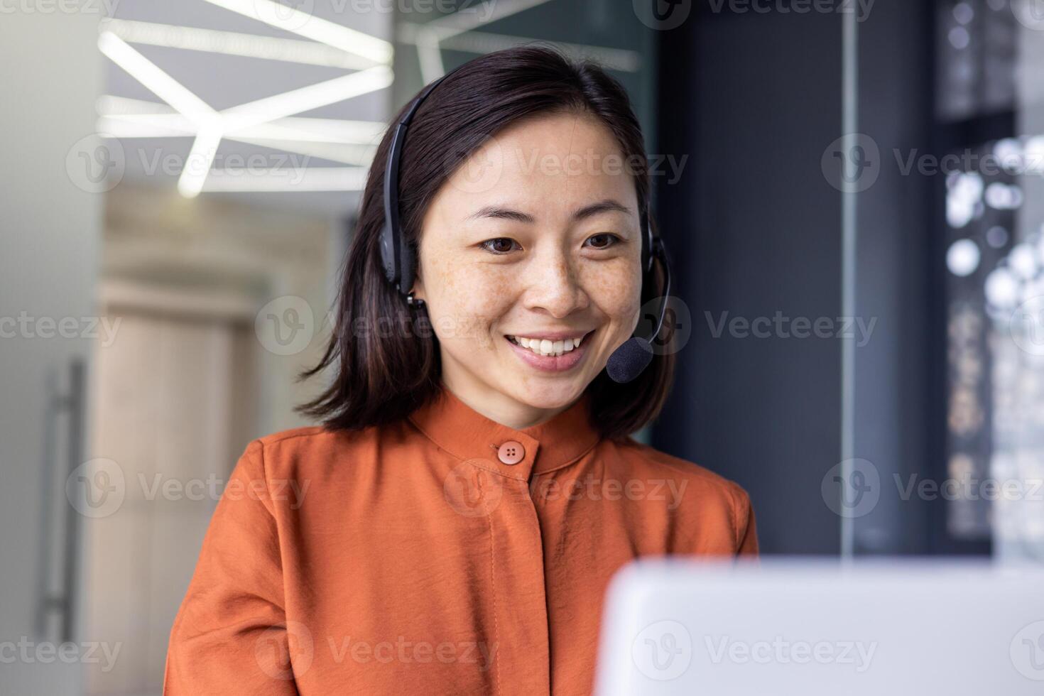 Close-up photo of young beautiful Asian woman talking on call, looking at laptop web camera and smiling using headset for communication, successful businesswoman working in office.