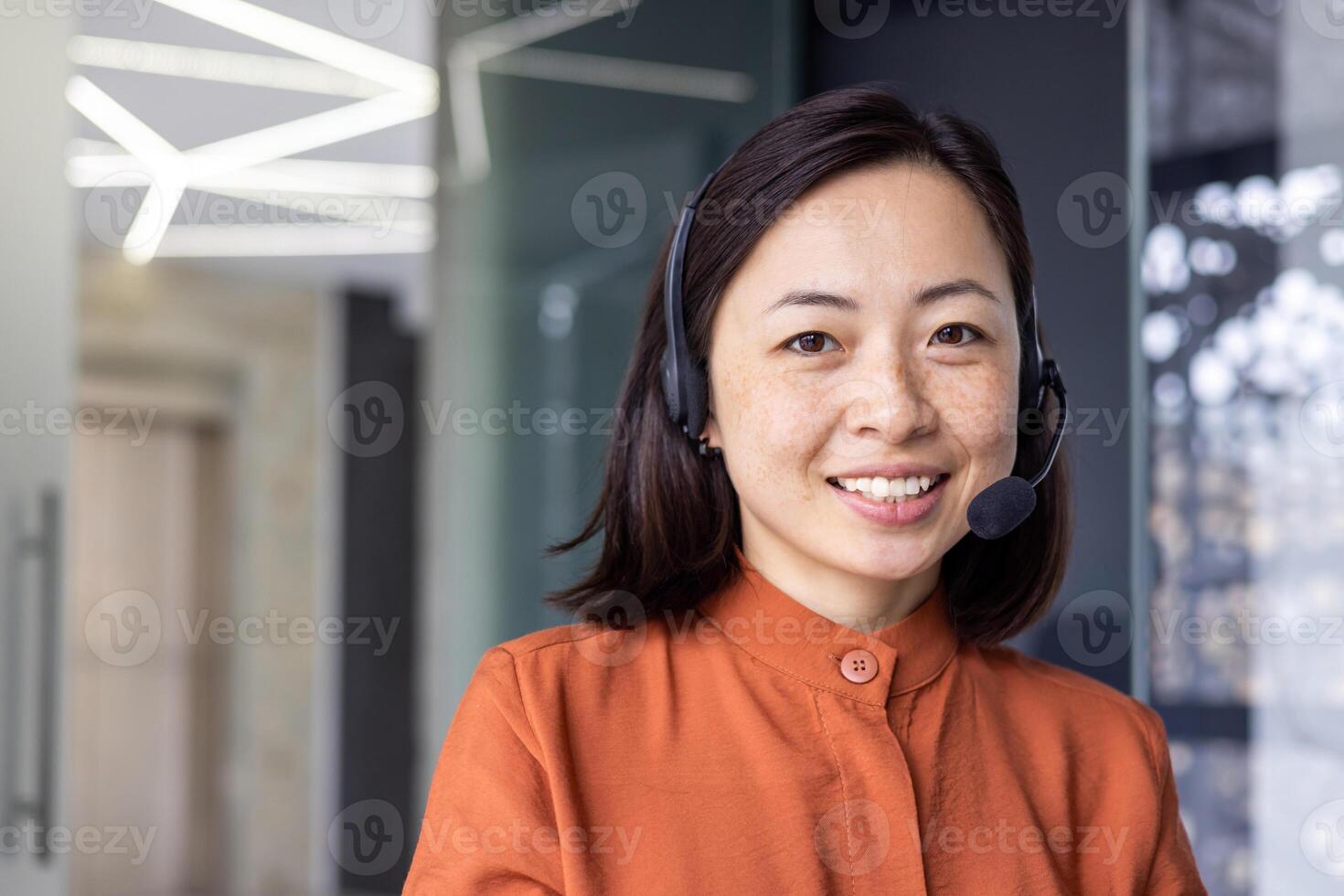 Close up portrait of Asian woman inside modern office with headset for call, woman smiling and looking at camera, office worker customer support tech helpline. photo