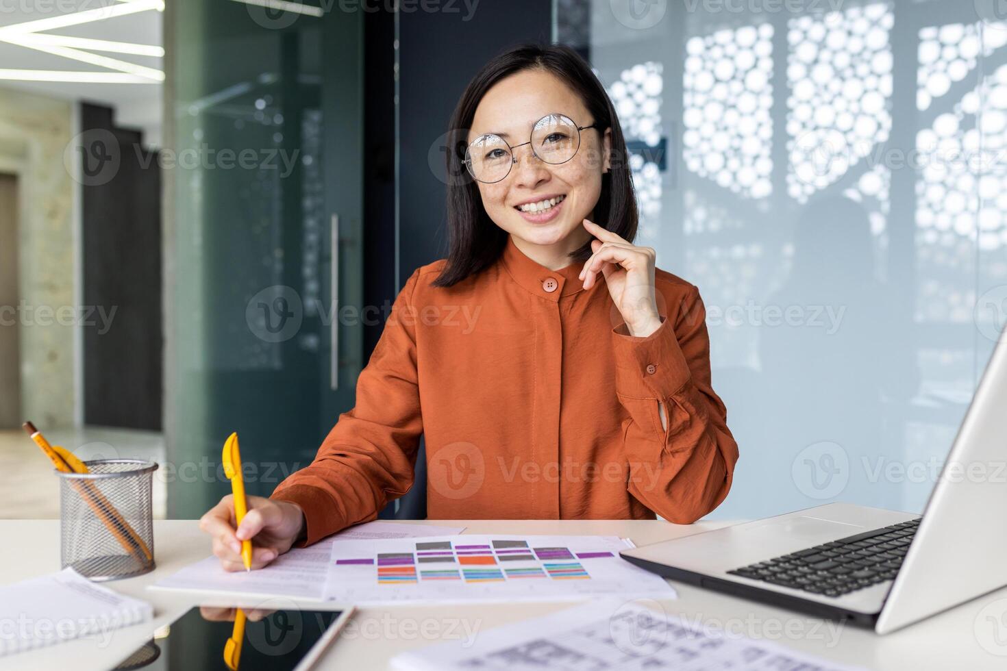 Portrait of successful Asian financier doing paperwork, businesswoman smiling and looking at camera, filling out documents, forms at workplace, writing documents inside office, using laptop. photo