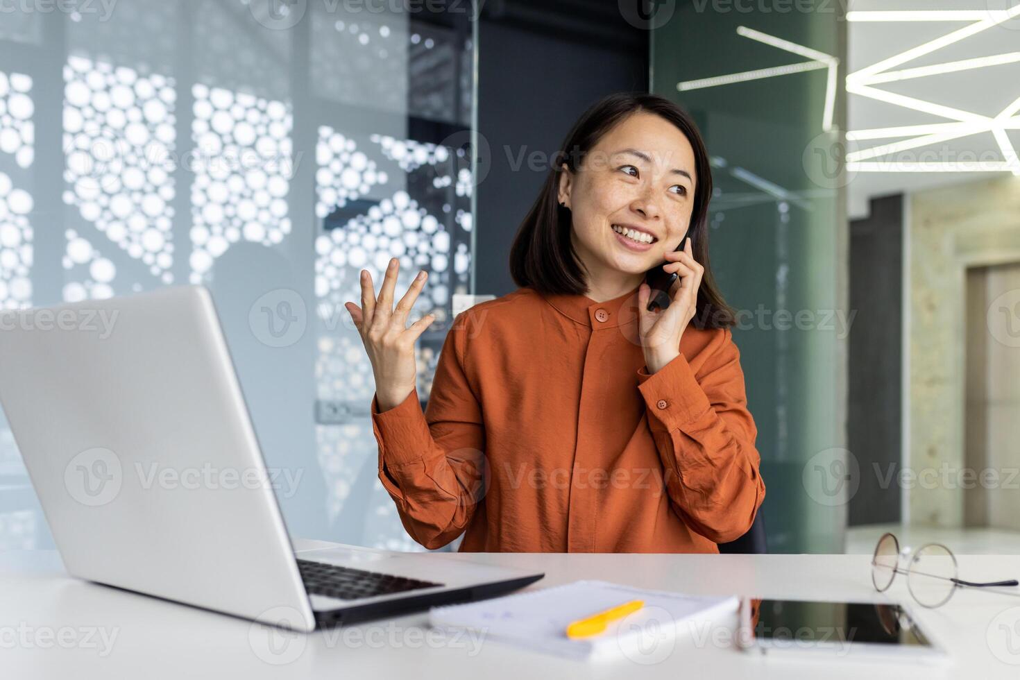 Young successful Asian businesswoman working inside modern office, female employee cheerfully talking to colleagues on phone and smiling, using laptop while sitting at desk. photo