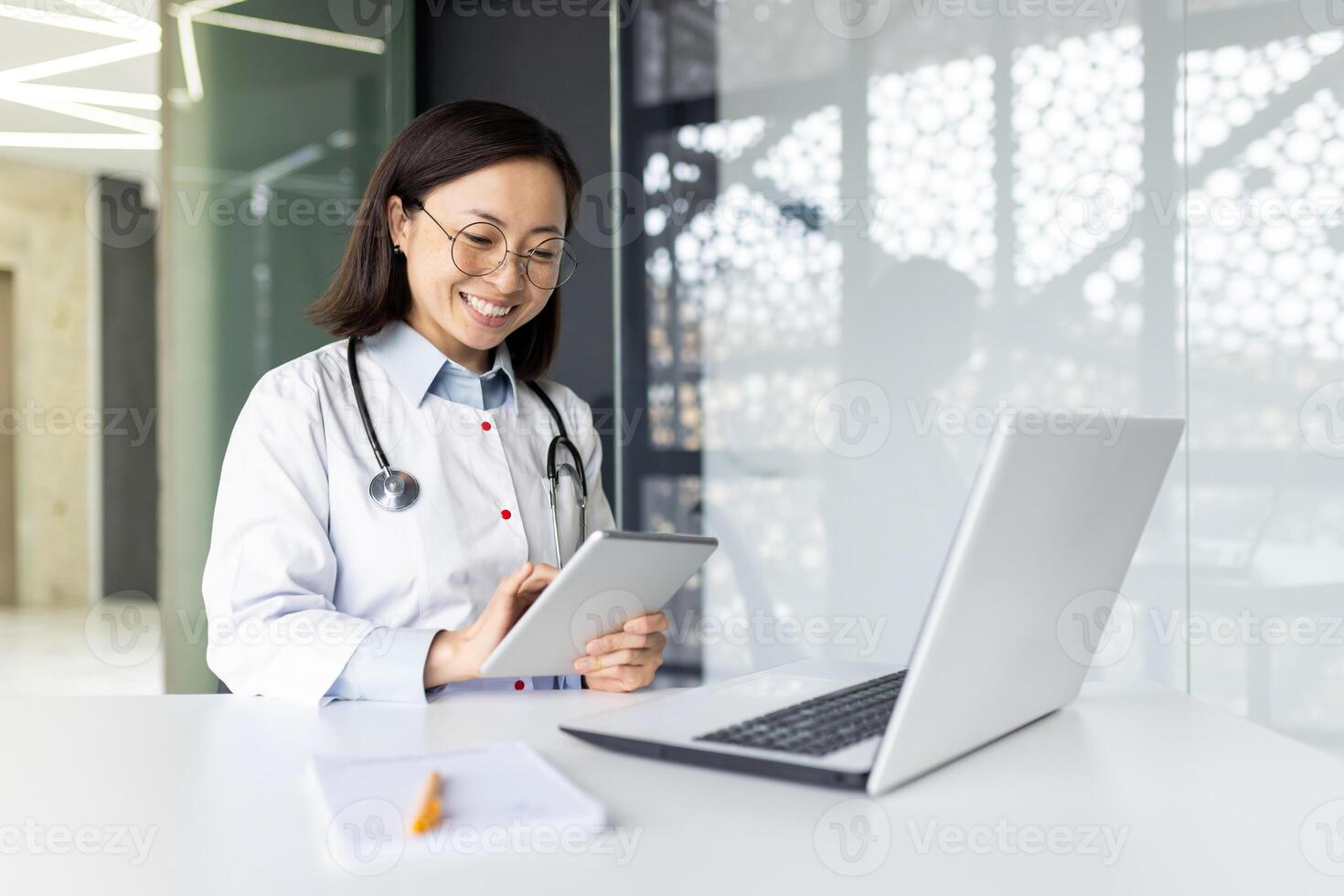 Young smiling Asian woman doctor, student sitting in the hospital office at the work table in a white coat, working on studying on a laptop, making notes in a notebook. photo