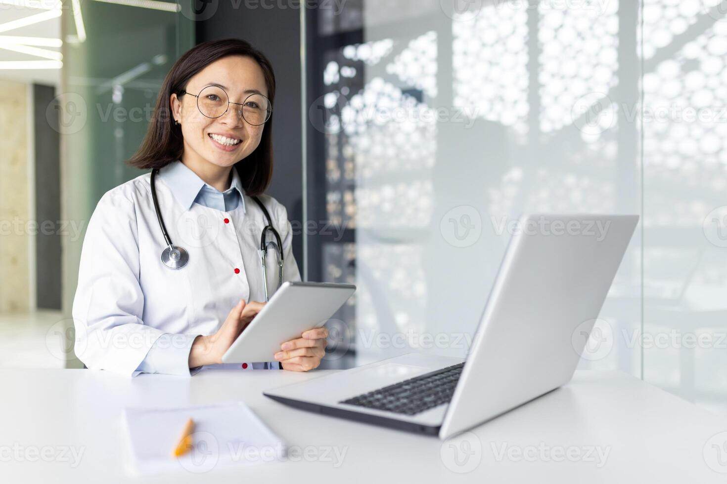 Joyful asian intern woman dressed in medical coat sitting at desktop with laptop and holding digital tablet in hands. Smiling female nurse using modern gadgets for productive work in hospital office. photo