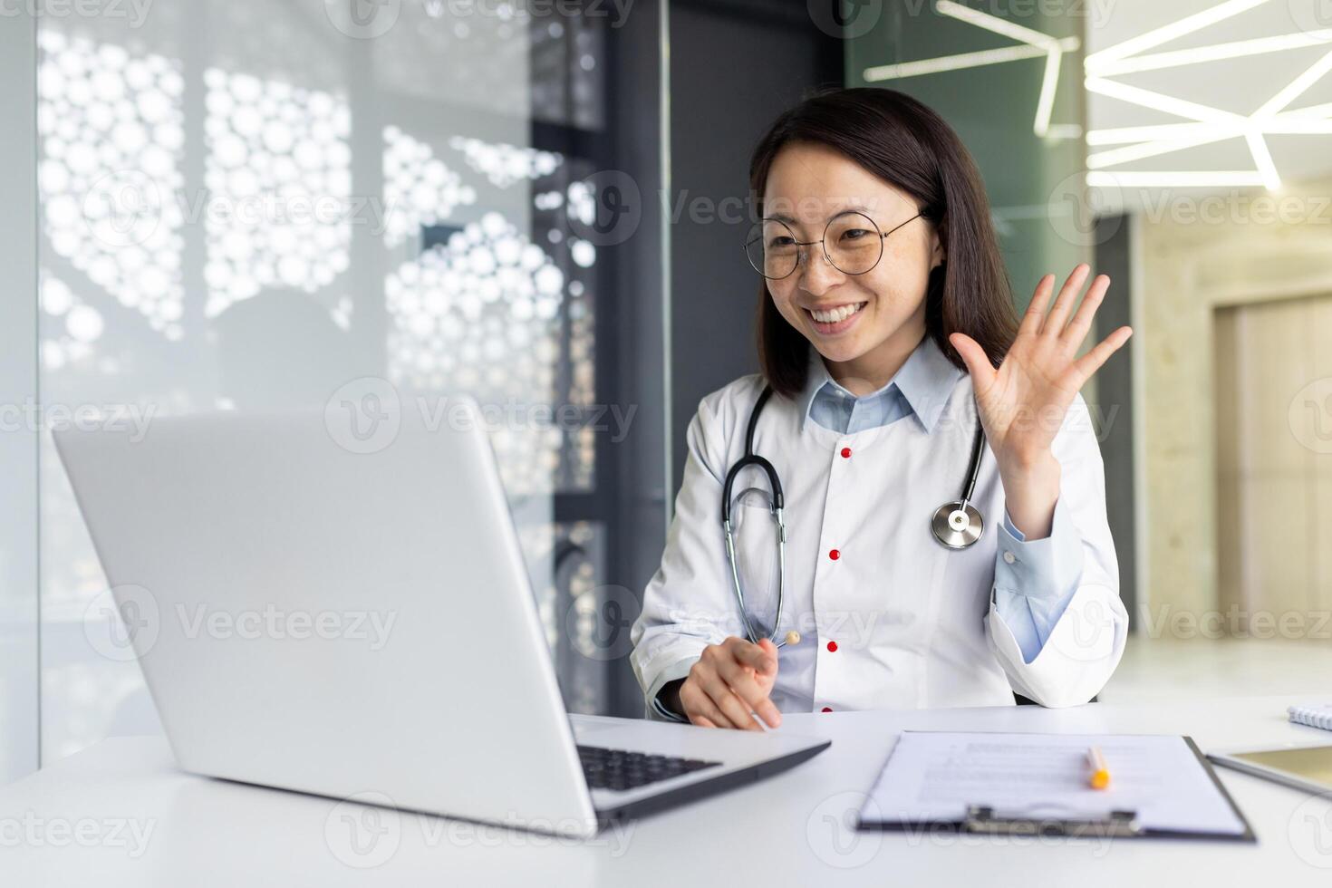 Smiling Asian young female doctor consulting online patient. Sitting at a table in a hospital office in a white coat, talking and greeting on a call on a laptop. photo