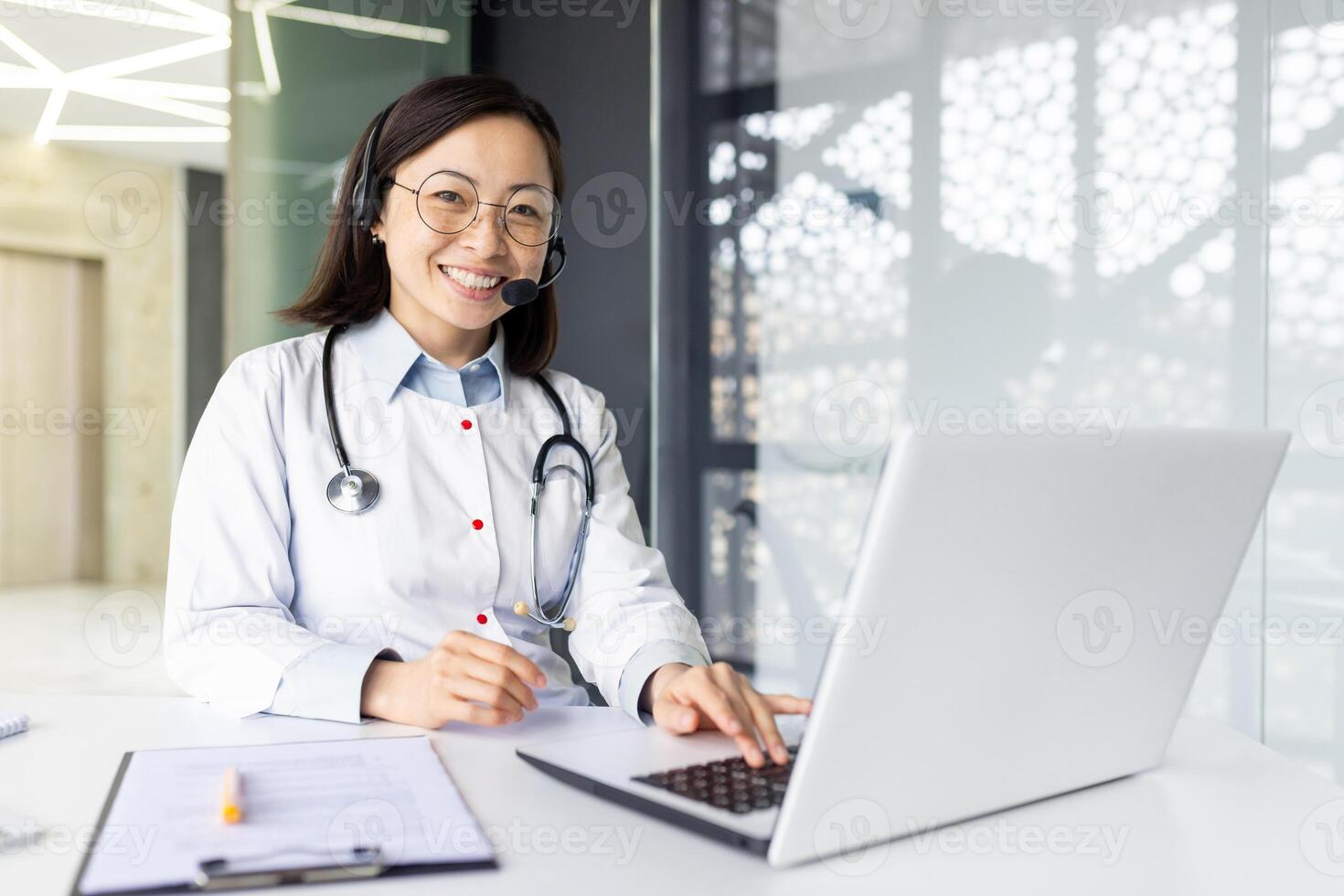 Portrait of a smiling young female Asian doctor sitting in the clinic at the work table in a headset and white coat in front of a laptop, working remotely, looking confidently into the camera. photo